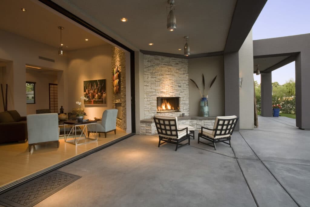 Elegant indoor-outdoor living space with sliding glass doors, stone fireplace, modern furniture, and smooth concrete floor under open sky.